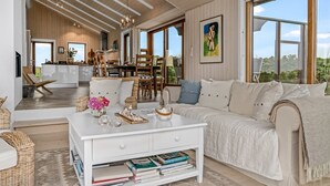 Amplia sala de estar con un sofá grande, una mesa de centro con libros y vistas al exterior a través de amplios ventanales.