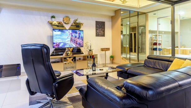 A modern living room with a black leather sofa, a glass coffee table, a black office chair, a flat-screen TV, and a decorative wall piece. - Xspa apartment cryptocurrency pay available (Novi Sad)