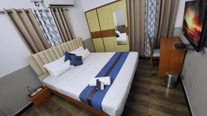 A hotel room with a bed, a wooden nightstand, a desk, a trash bin, and a TV on the wall. - Ashirwad Residency  (Bengaluru)