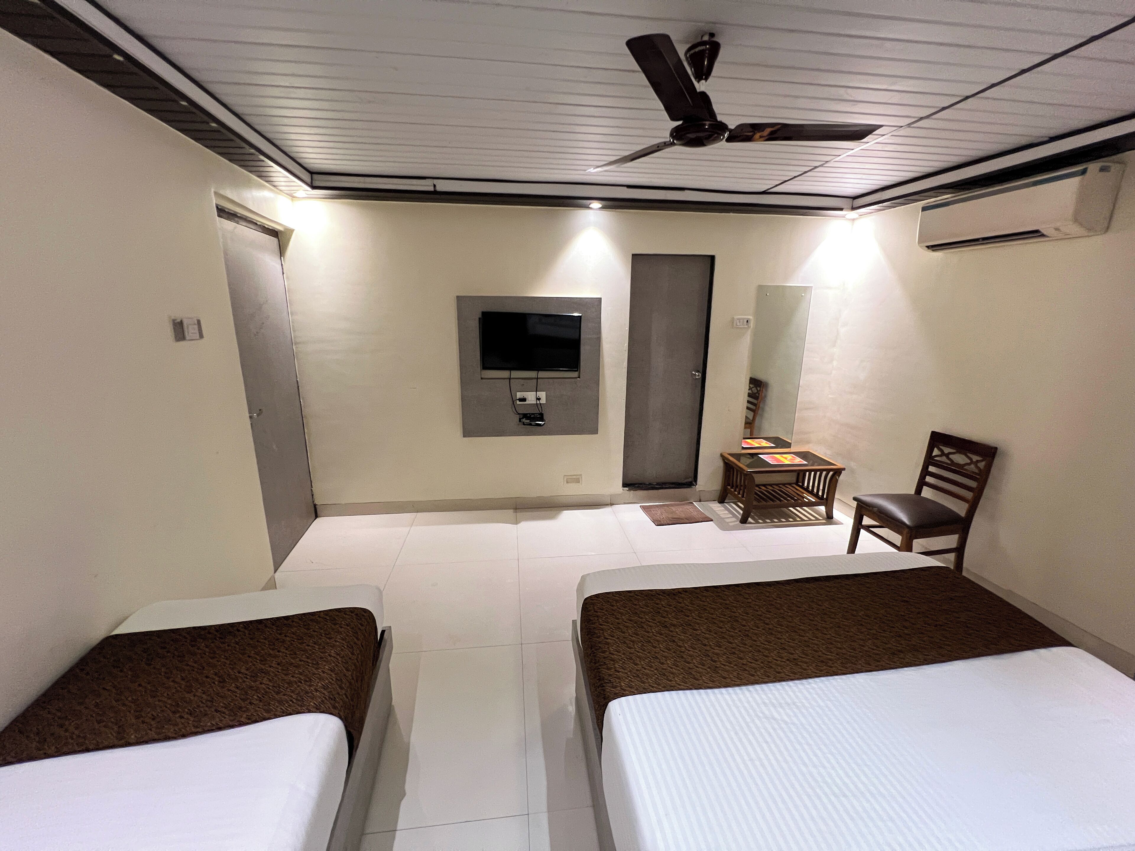 Une chambre d’hôtel avec deux lits, un téléviseur fixé au mur, un ventilateur de plafond et une petite table avec une chaise.