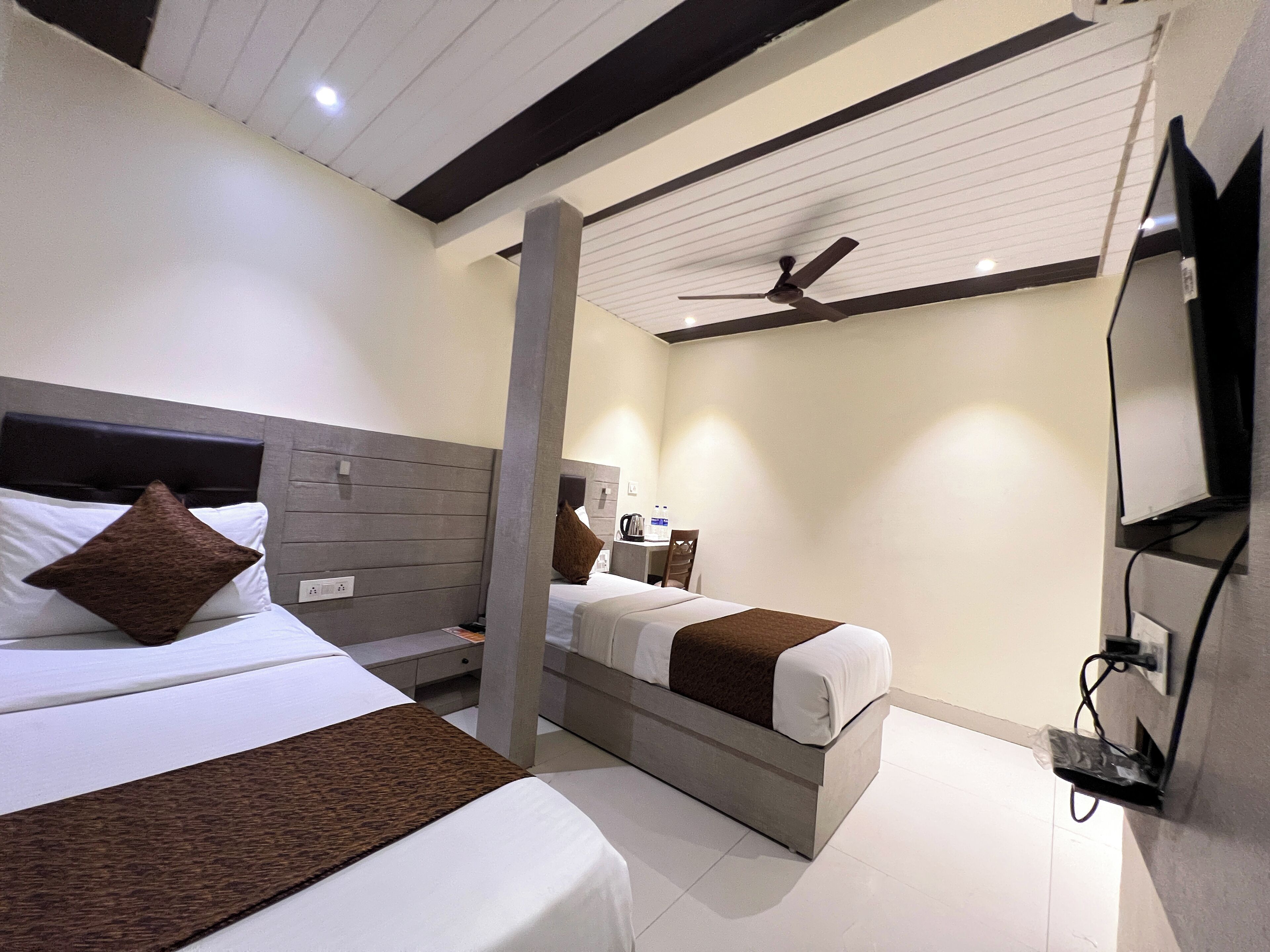Habitación de hotel con dos camas, un televisor montado en la pared, un ventilador de techo y una mesita de noche con una lámpara y una cafetera.