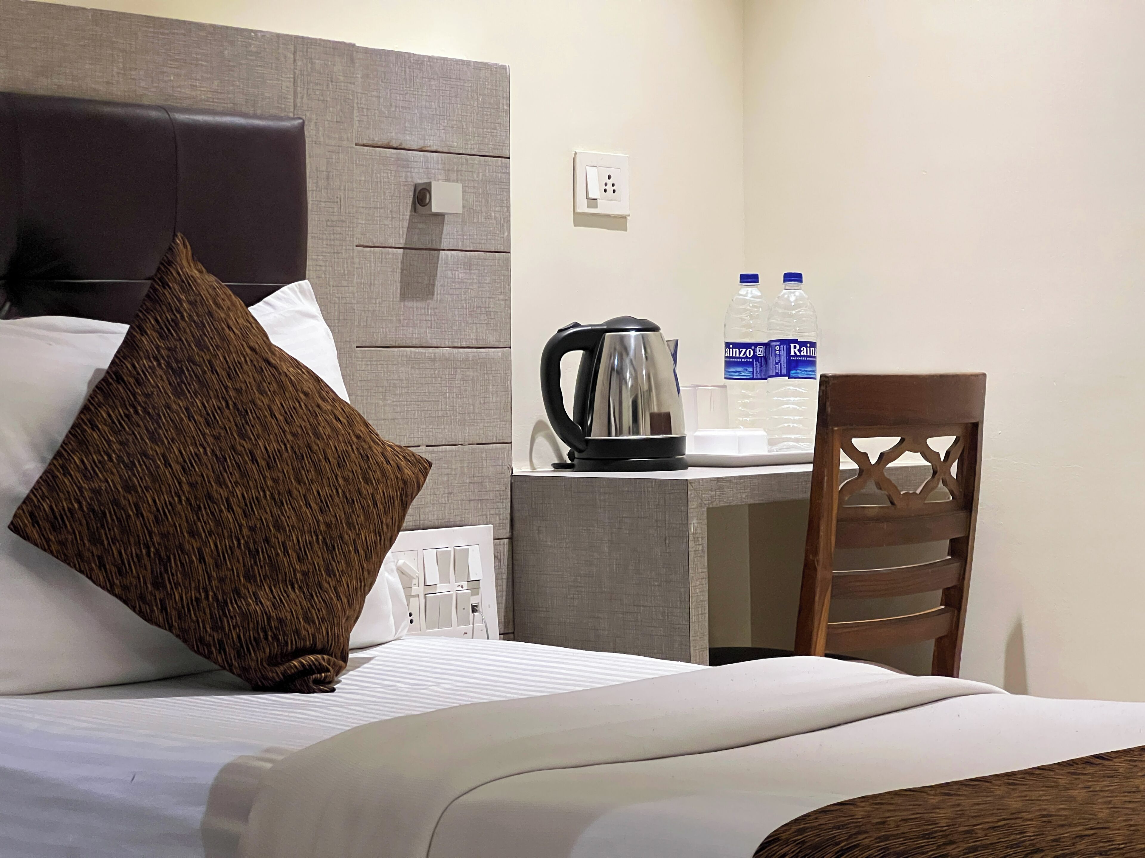 Quarto de hotel com cama, escrivaninha, cadeira, chaleira e duas garrafas de água.