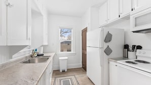 A compact kitchen with white cabinets, a stainless steel sink, a refrigerator, and a stove. - Charming Centrally Located Sunroom/Fireplace (Sioux Falls)