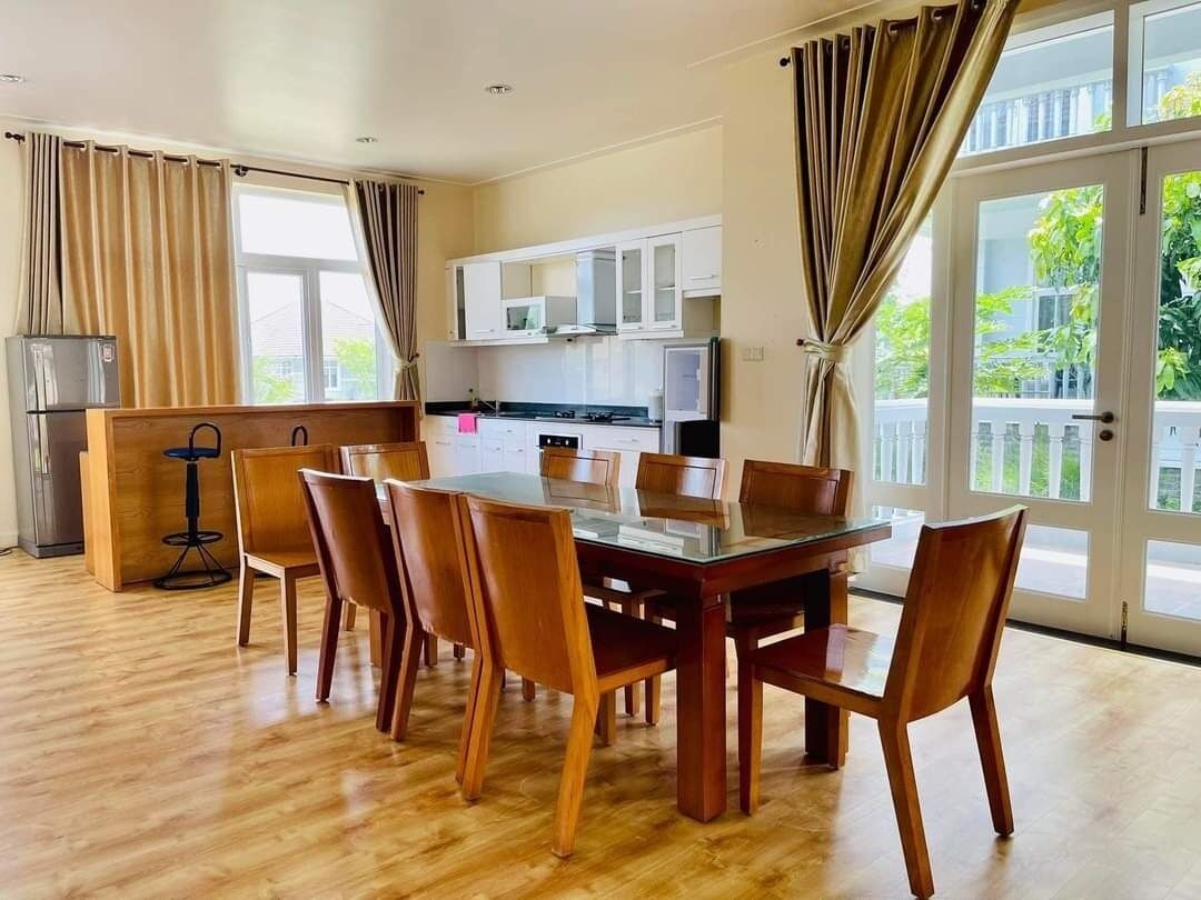 Un comedor amplio con una mesa y sillas de madera, una cocina con gabinetes blancos y ventanas grandes con cortinas.