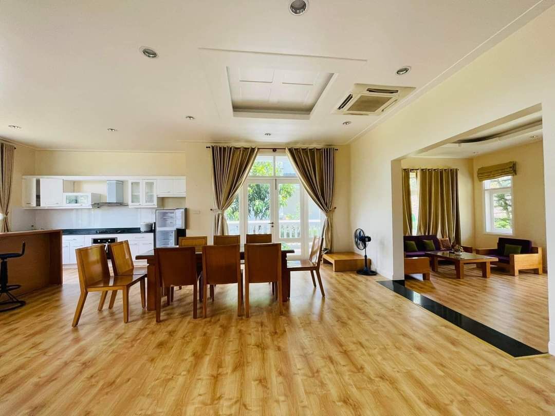 Uma sala de estar ampla com uma mesa de jantar e cadeiras, uma cozinha ao fundo e grandes janelas com cortinas.
