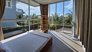 Une chambre avec un grand lit, une chaise en bois, un ventilateur sur pied et une vue sur un balcon donnant sur une piscine et un espace verdoyant.