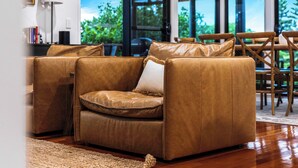 A modern living room with a brown leather sofa, wooden flooring, and large windows offering a view of greenery.