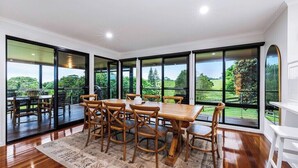 A dining area with a wooden table and chairs, large windows, and a view of greenery.