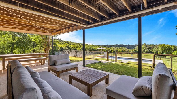 An outdoor patio with a wooden ceiling, cushioned seating, and a view of a green landscape.