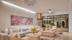 Ein modernes Wohnzimmer mit einer Couch, einem Couchtisch und Sesseln. An der Wand hängt ein großes abstraktes Gemälde und eine Glastür führt in einen weiteren Raum.
