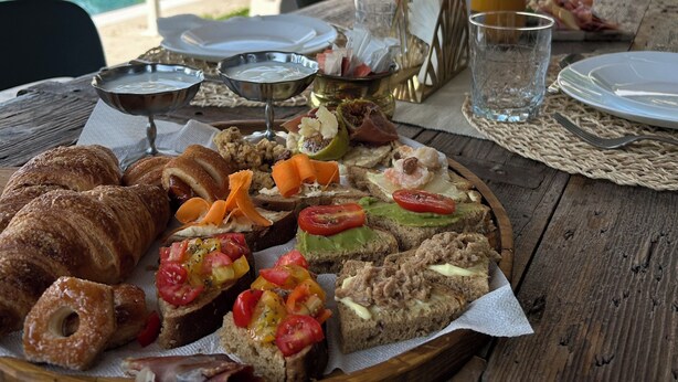Buffet de café da manhã com croissants, sanduíches e frutas em uma mesa de madeira ao lado de uma piscina.