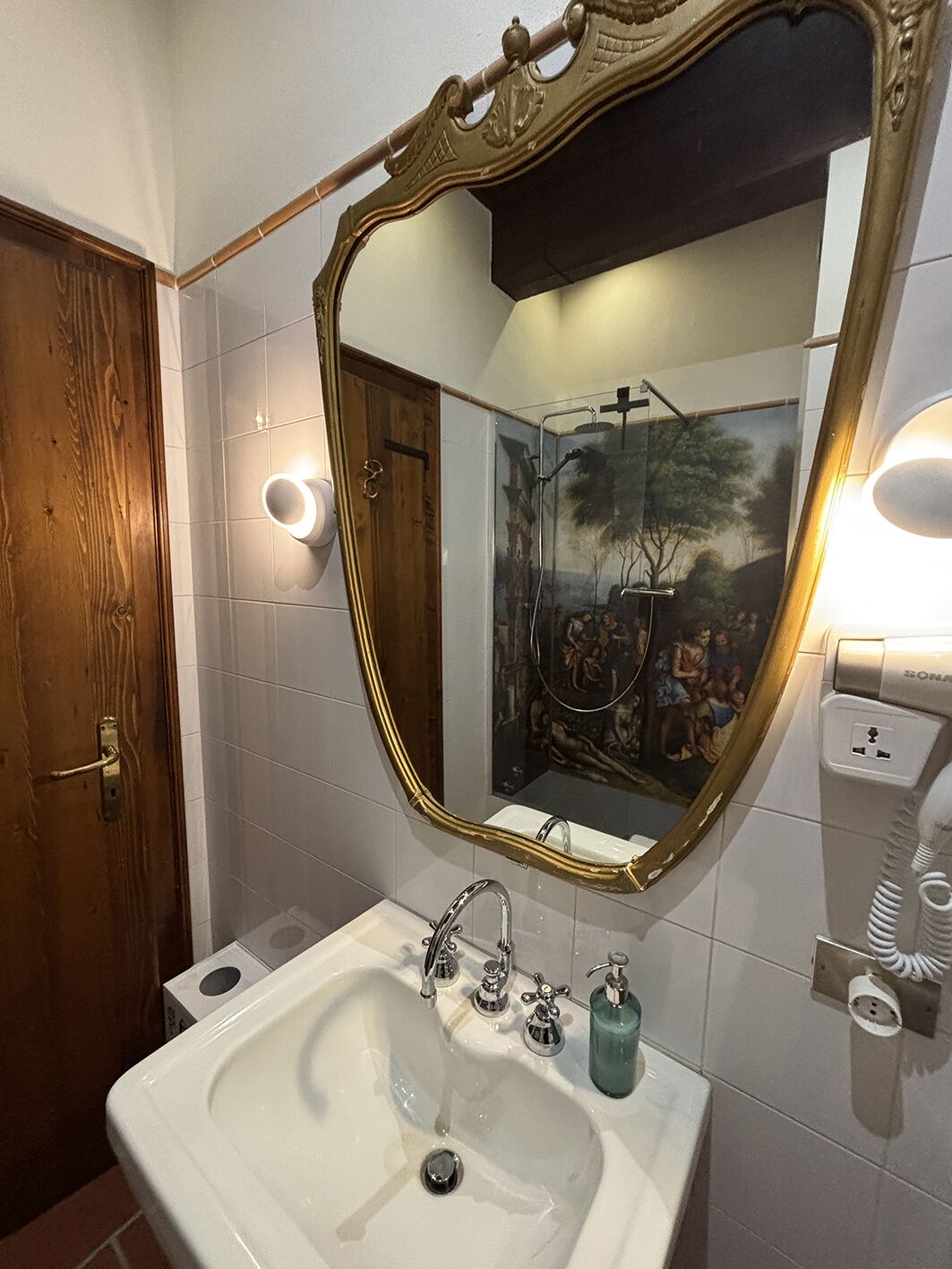 Une salle de bain dotée d’un grand miroir décoratif, d’un lavabo blanc avec plusieurs robinets et d’un luminaire fixé au mur.