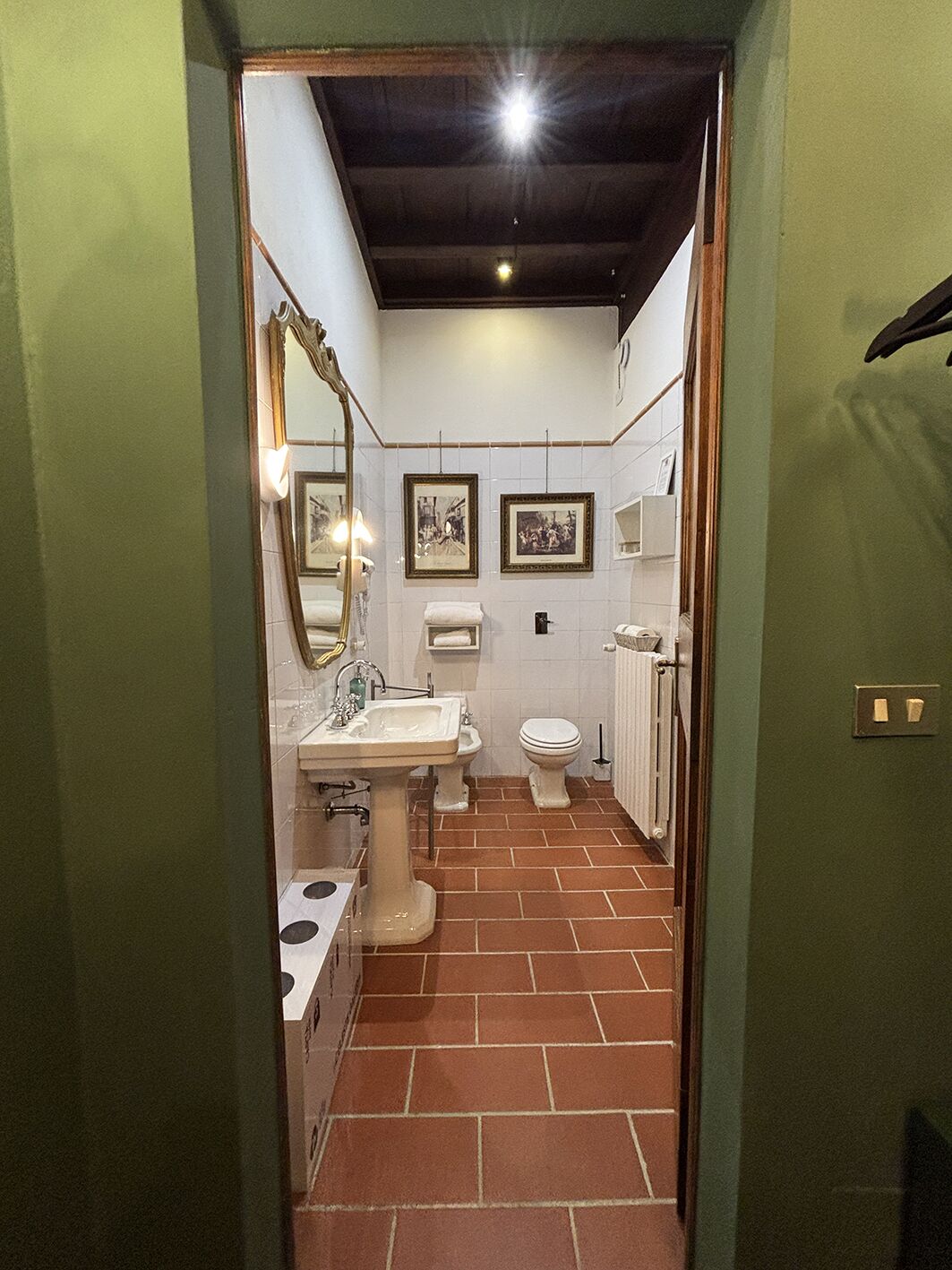 A narrow, tiled bathroom with a pedestal sink, a toilet, and a mirror.