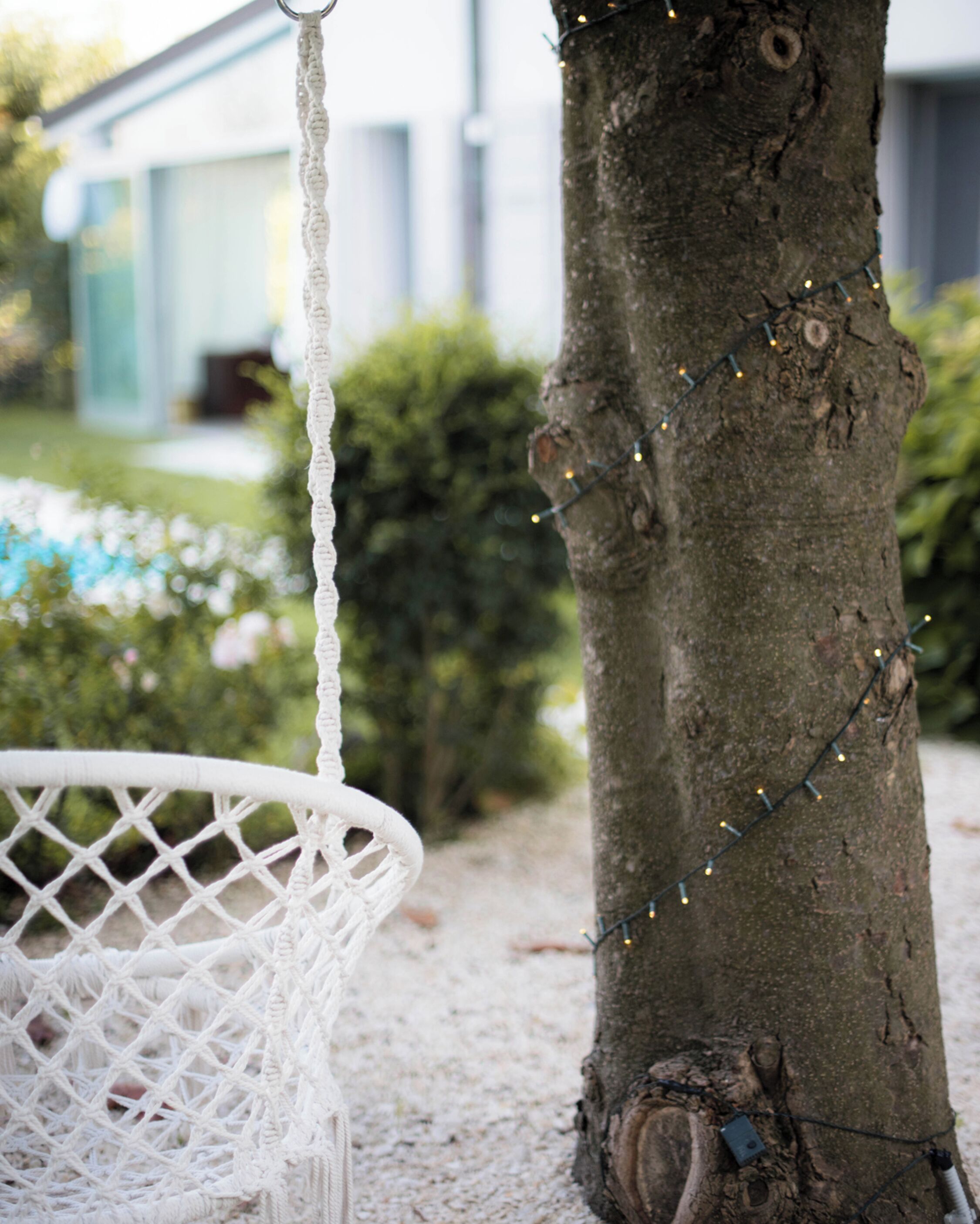 A white wicker chair with a macrame hanging swing, a tree with string lights, and a building in the background.