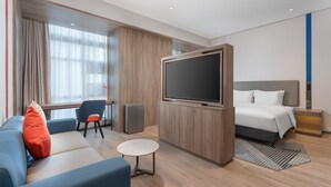 A modern hotel room with a large flat-screen TV, a sofa, a chair, a small round table, and a bed with a headboard. - Holiday Inn Express Guangzhou University Town by IHG (Guangzhou)