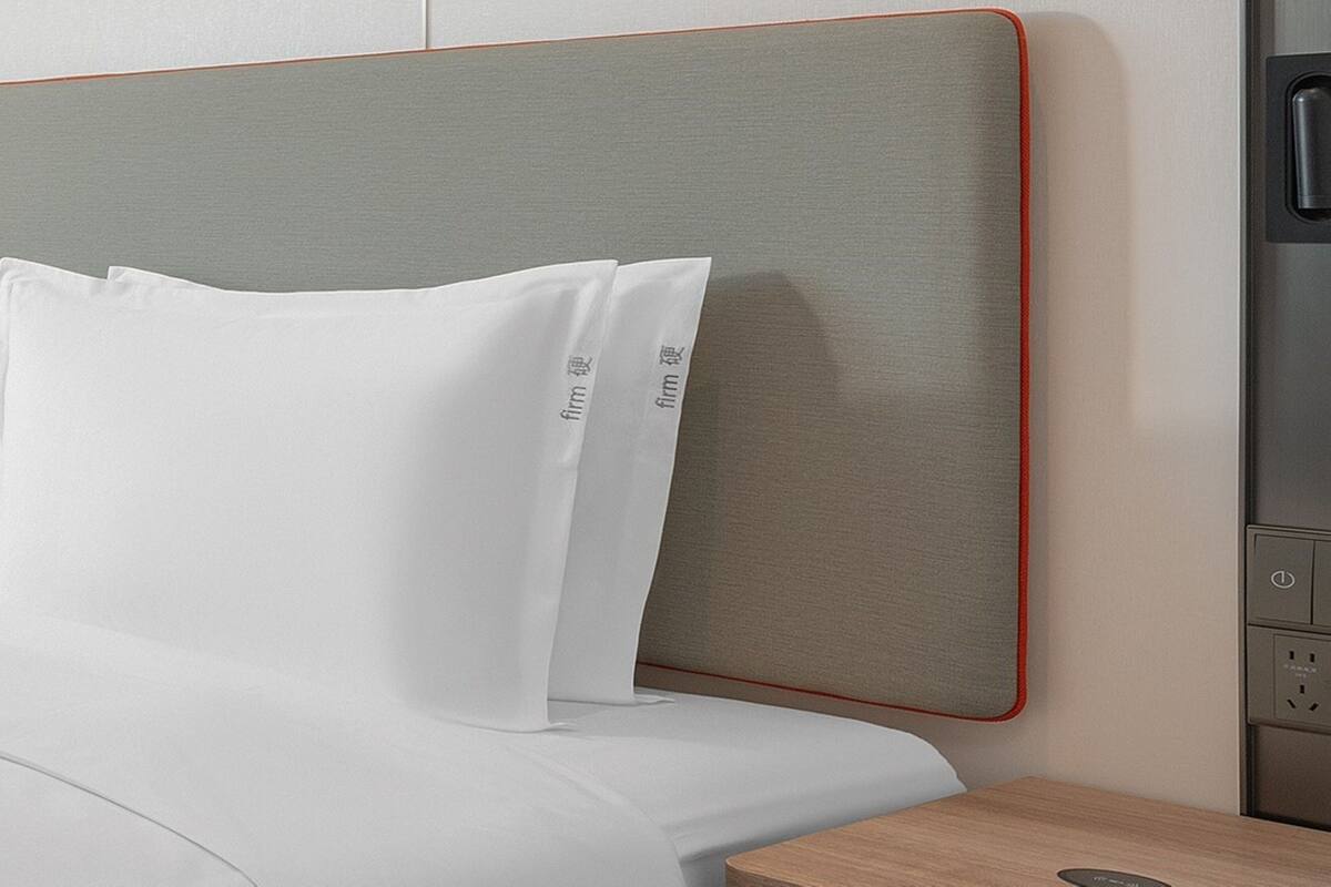 A neatly made bed with white linens and pillows, a grey headboard with a red trim, and a wooden bedside table with a power outlet.