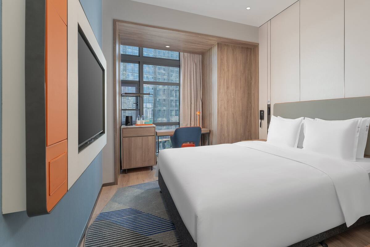 A hotel room with a large bed, a desk with a chair, a TV, and a window with a city view.