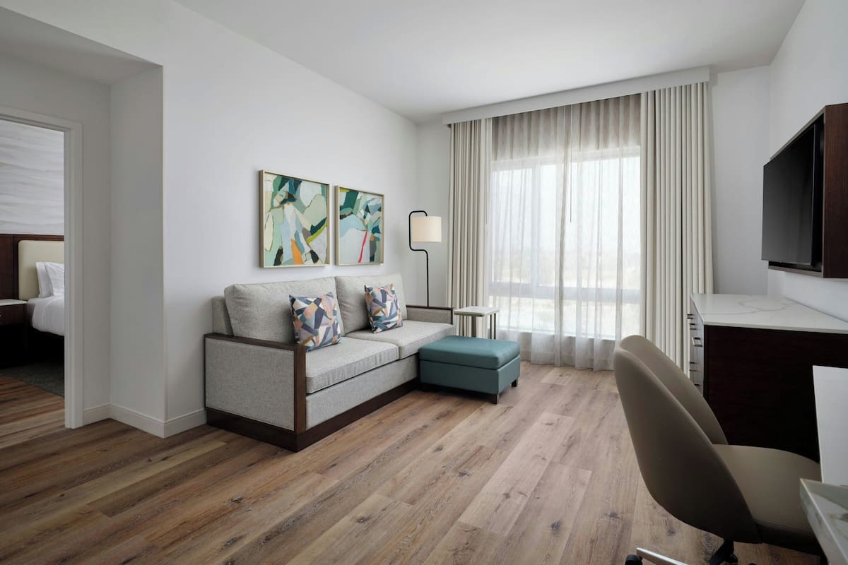 A modern hotel room with a sofa, ottoman, and a desk with a chair. There is a TV mounted on the wall and a floor lamp.