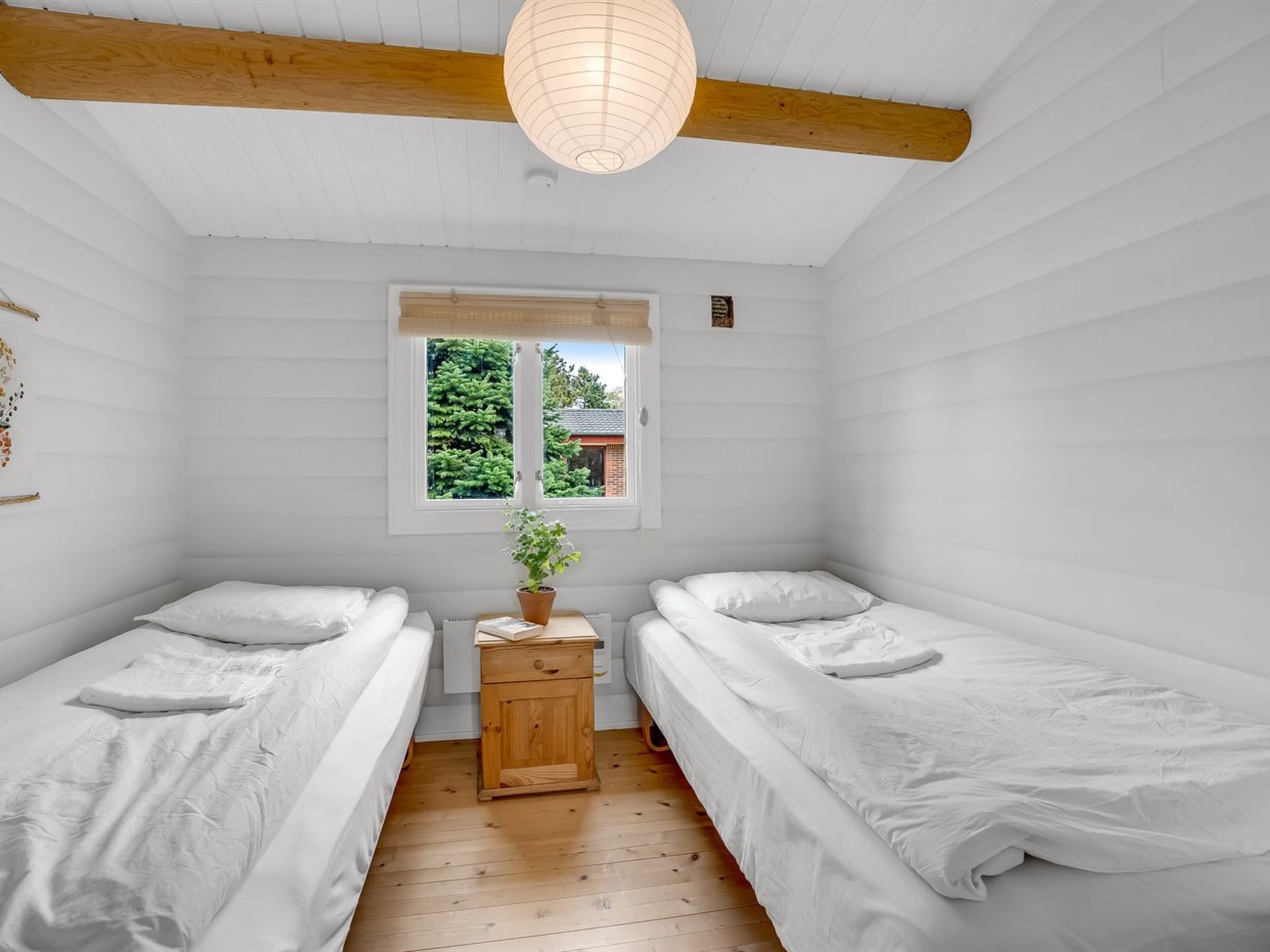 Une petite pièce aux murs recouverts de panneaux blancs, avec deux lits simples, une table de chevet en bois ornée d’une plante, et une fenêtre donnant sur un paysage verdoyant.