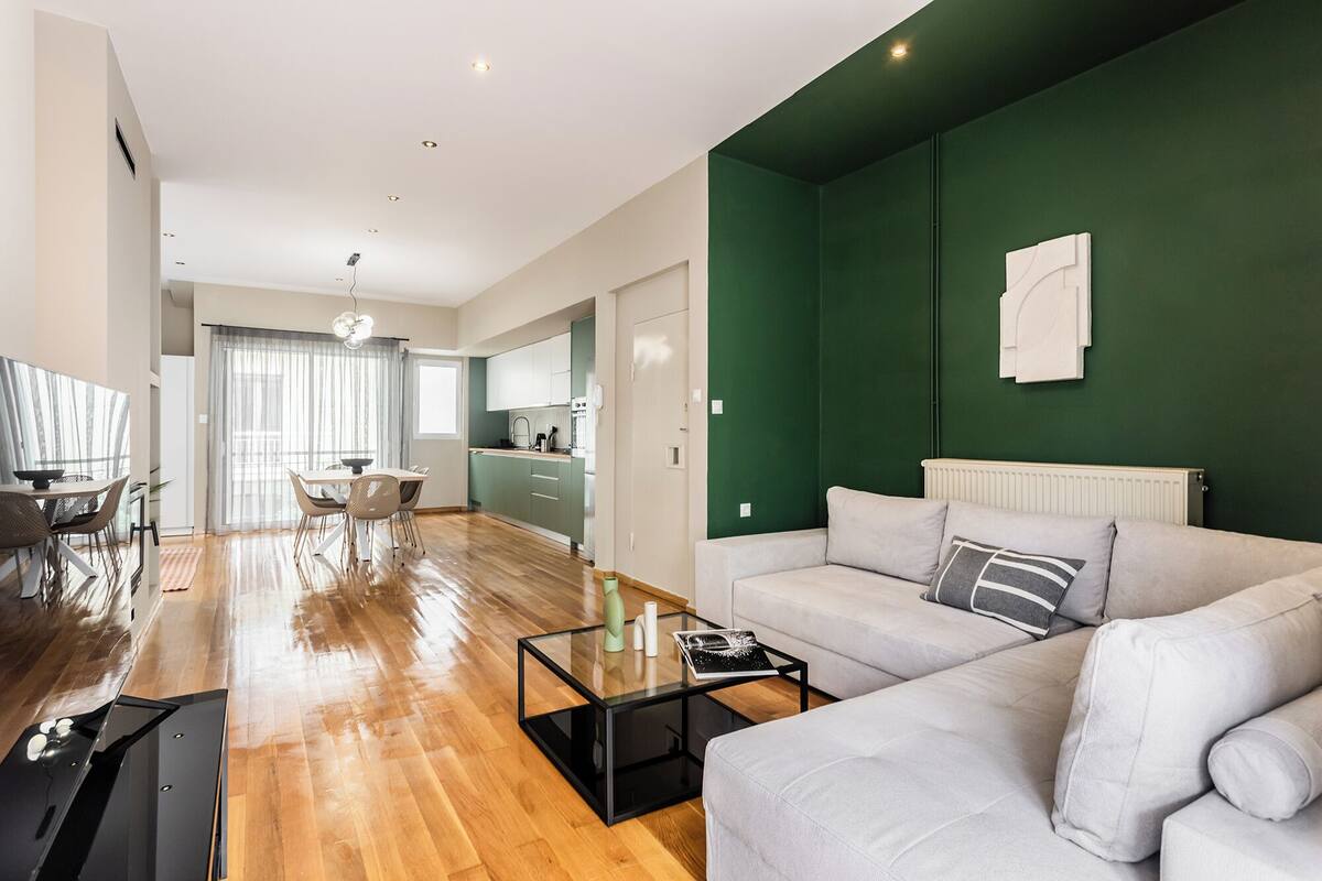 A modern living room with a green accent wall, a sofa, a glass coffee table, and a dining area with a table and chairs.