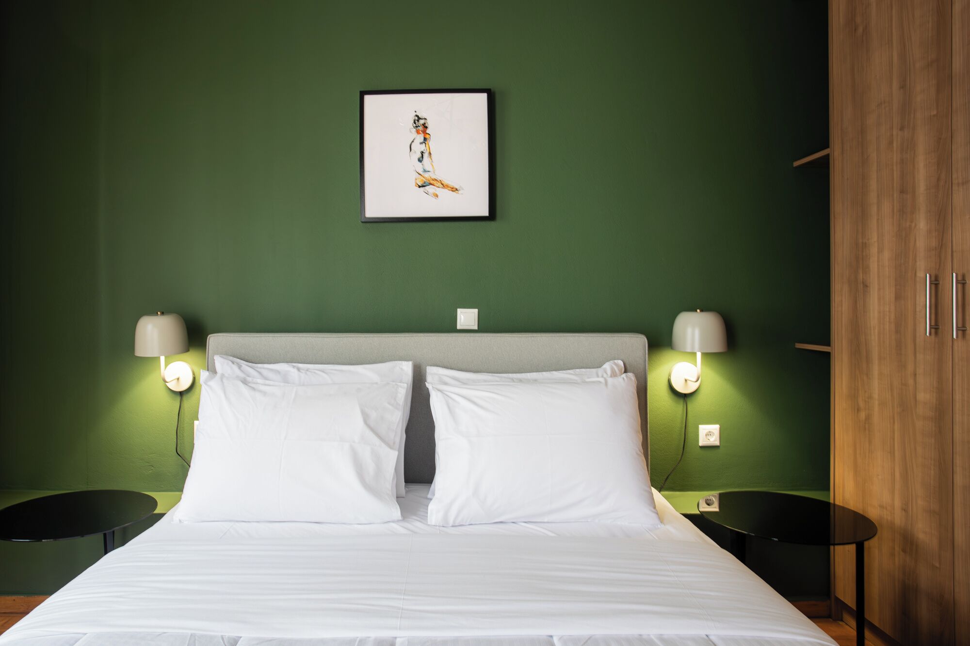 Quarto de hotel com cama, duas mesas de cabeceira, um criado-mudo, parede verde, quadro emoldurado e guarda-roupa de madeira.