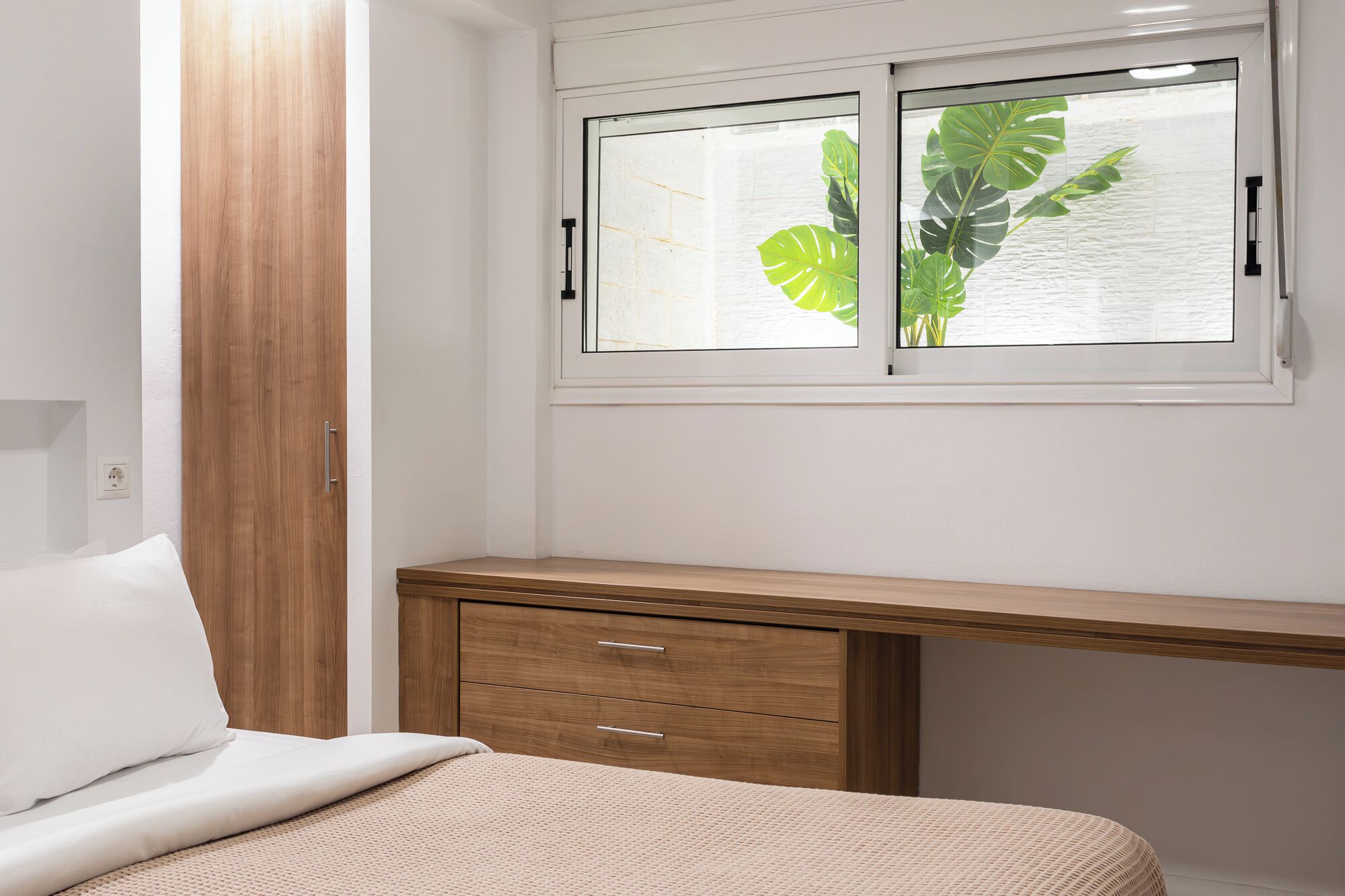 A bedroom with a bed, a wooden desk, and a window with a view of a plant.