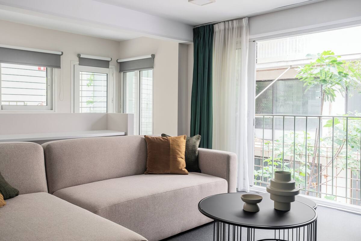 A modern living room with a beige sofa, a round black coffee table, and a view of a balcony with greenery.