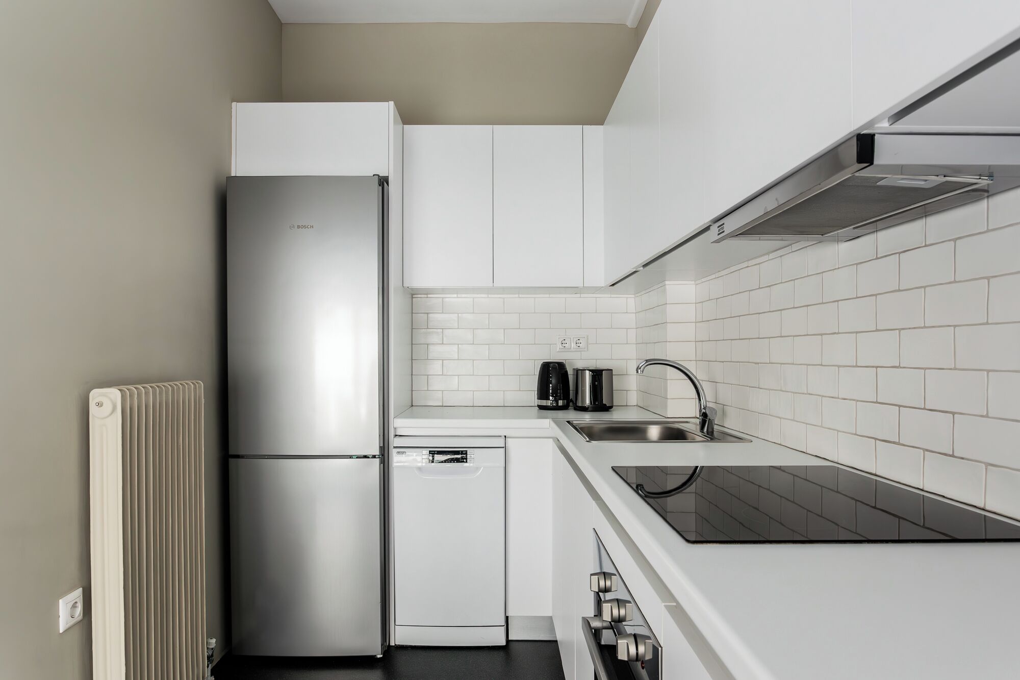 Una cucina compatta con mobili bianchi, un frigorifero in acciaio inossidabile, una lavastoviglie, un piano di lavoro nero e un lavello con un rubinetto moderno.