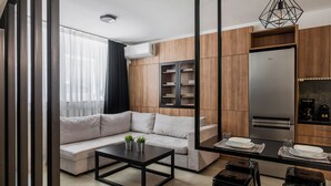 Una sala de estar moderna con un sofá, una mesa de centro y una zona de comedor con taburetes. Hay una estantería y una cocina con un refrigerador y una cafetera.