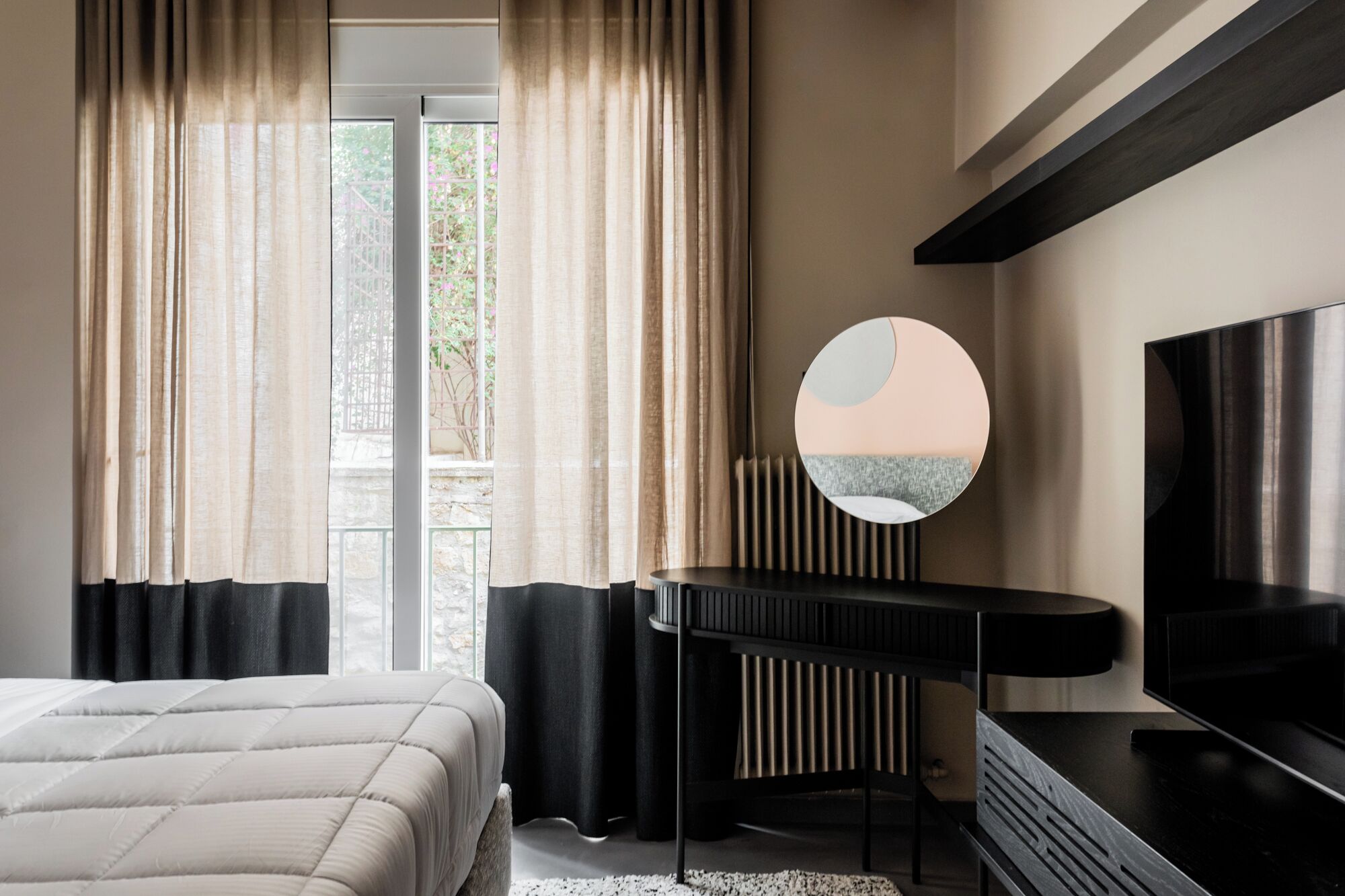 Quarto moderno com cama, escrivaninha, espelho redondo e televisão.
