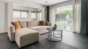 Un salón moderno con un sofá seccional color beige, una mesa de centro redonda y vistas a un balcón con barandilla y follaje.