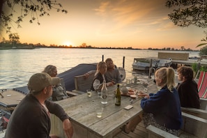A group of people seated at a wooden dock table with drinks, overlooking a body of water during sunset. - Pean Huur je Eigen Schier Eiland (Nes)