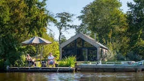 A riverside dock with a wooden structure, a boat, and a person sitting under an umbrella. - Pean Huur je Eigen Schier Eiland (Nes)