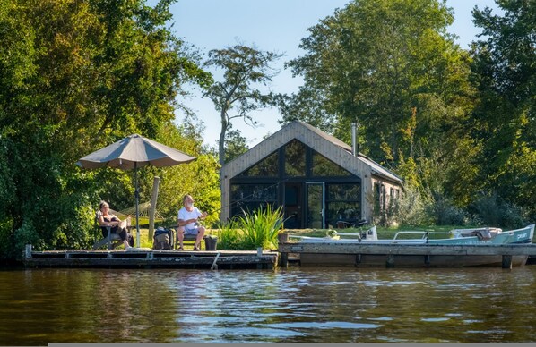 A riverside dock with a wooden structure, a boat, and a person sitting under an umbrella. - Pean Huur je Eigen Schier Eiland (Nes)