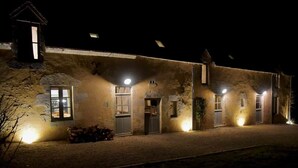 A rustic stone building with multiple windows and doors, illuminated by exterior lights. - To complete (Berd'huis)