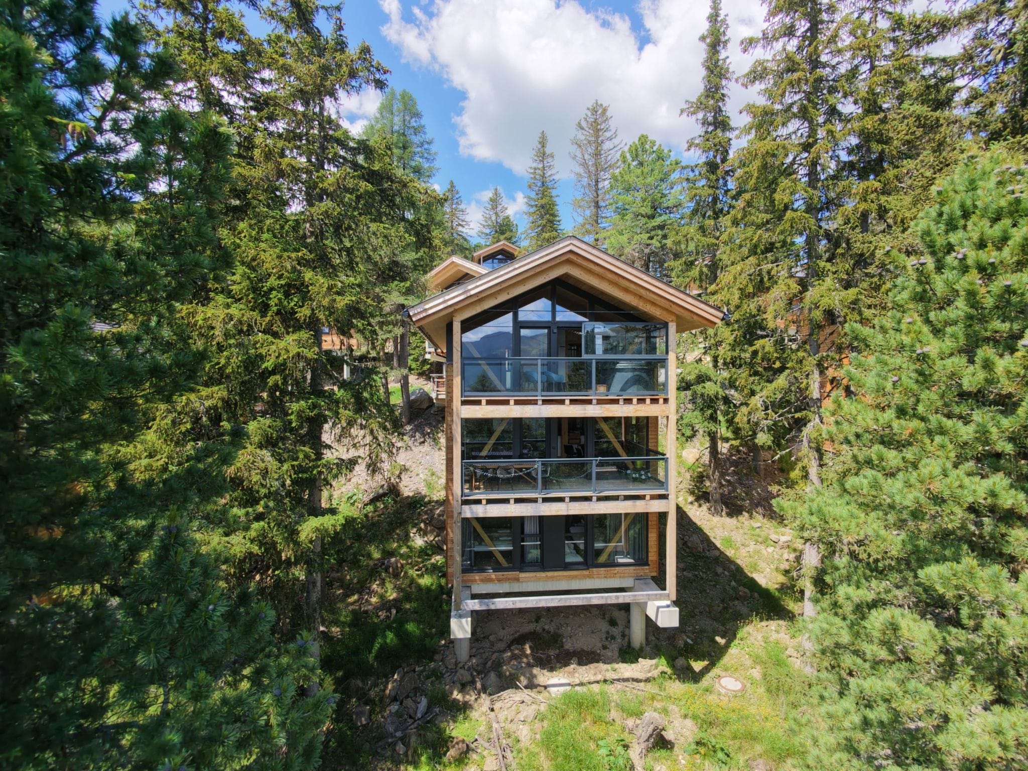 A two-story wooden cabin with large windows, surrounded by dense forest.