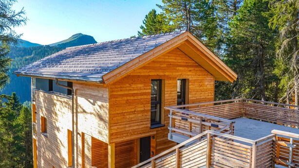 A wooden cabin with a balcony, set against a mountainous backdrop.