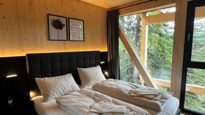 A bedroom with a large bed, wooden ceiling, and a view of trees through a window.