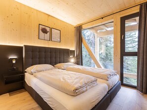 A bedroom with a wooden ceiling, a bed with two pillows, a bedside table with a lamp, and a window with curtains.