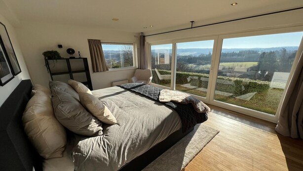 A bedroom with a large bed, a chair, a side table, and a view of the countryside through large windows. - Panoramic villa with private sauna in Erezée (Érezée)
