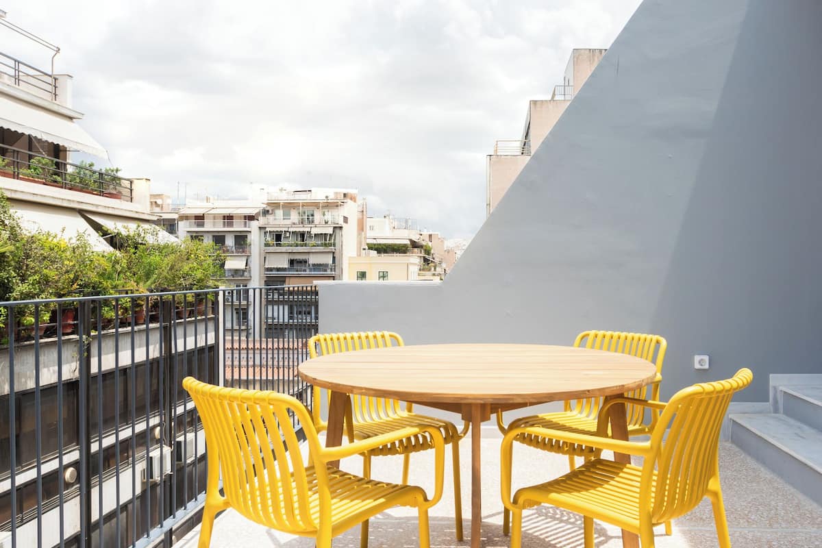 Una mesa redonda de madera con cuatro sillas amarillas en un balcón, con pared gris y barandilla de metal.