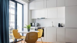 Cozinha moderna com armários brancos, forno embutido e cafeteira. Duas cadeiras de vime amarelas estão ao redor de uma mesa pequena com um vaso de planta.