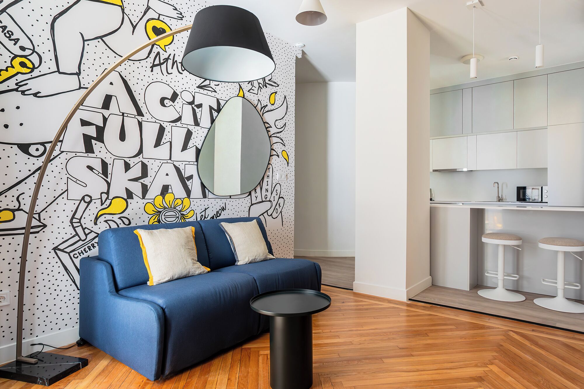 Una sala moderna con un sofá azul, una lámpara de pie negra y una mesa redonda negra. Se ve un área de cocina con gabinetes blancos y taburetes al fondo.