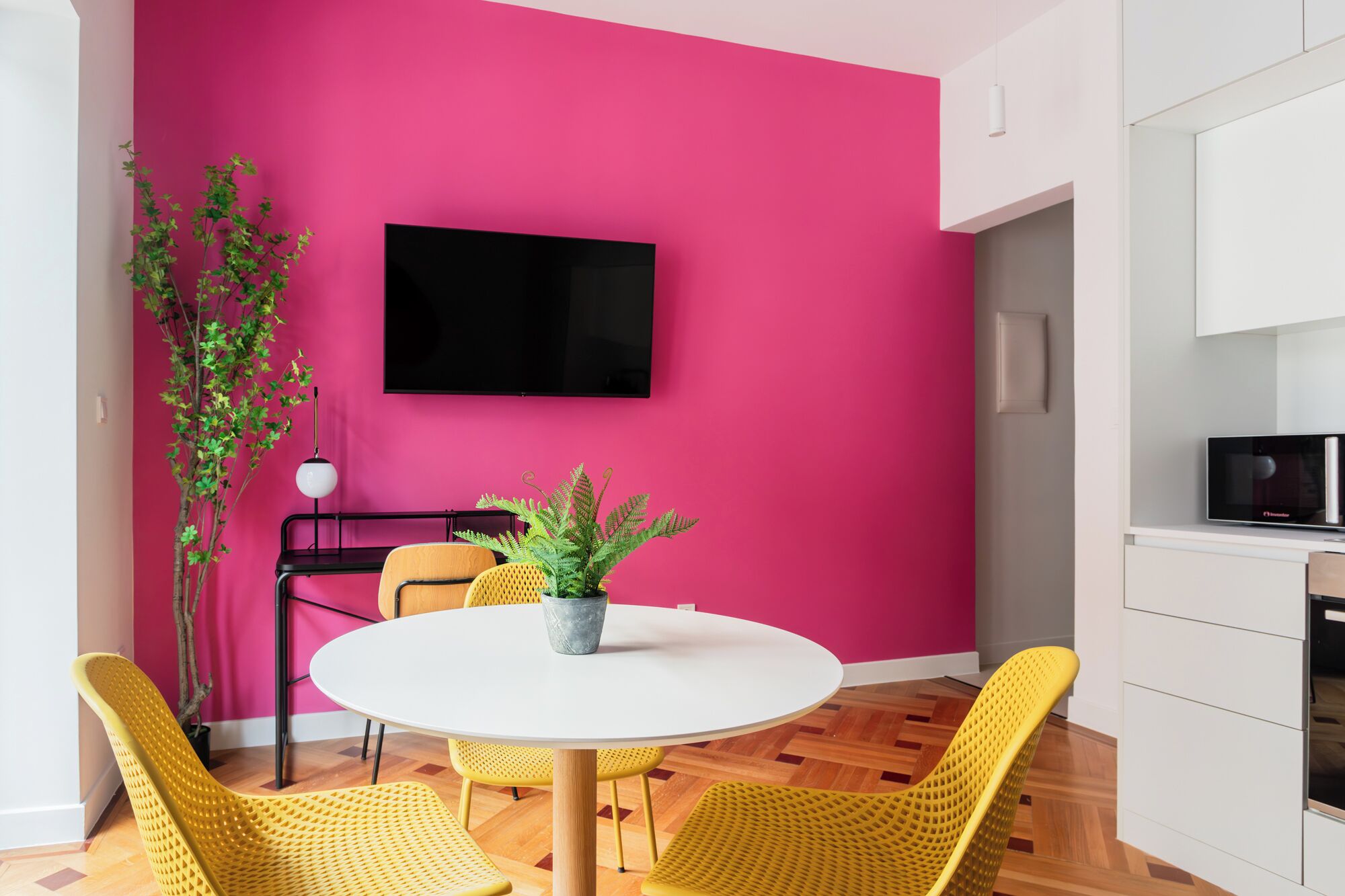 A modern kitchen with a pink wall, a flat-screen TV, and a dining area with yellow chairs and a white table.