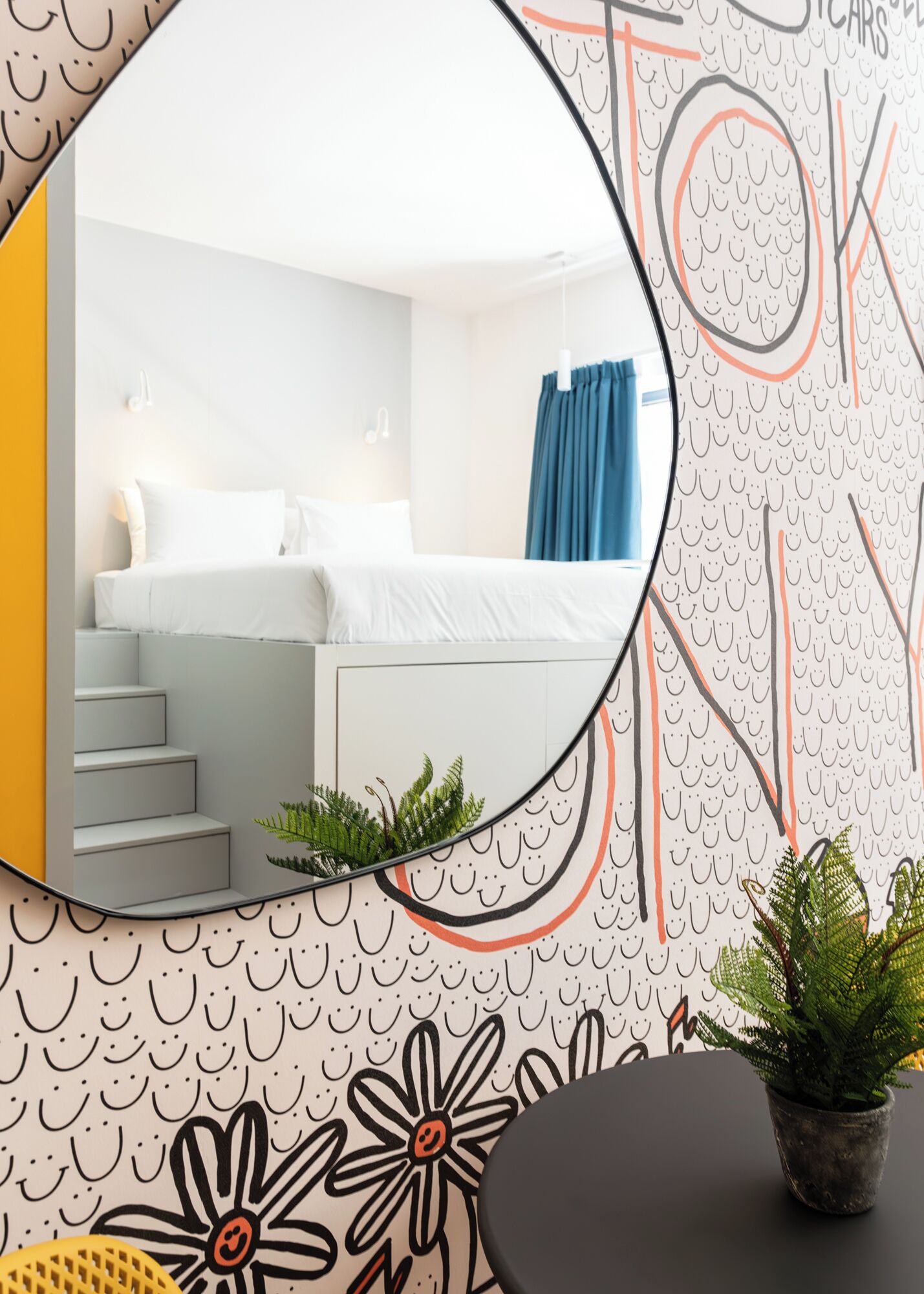 A modern hotel room with a bed, a small plant, and a mirror reflecting the room.