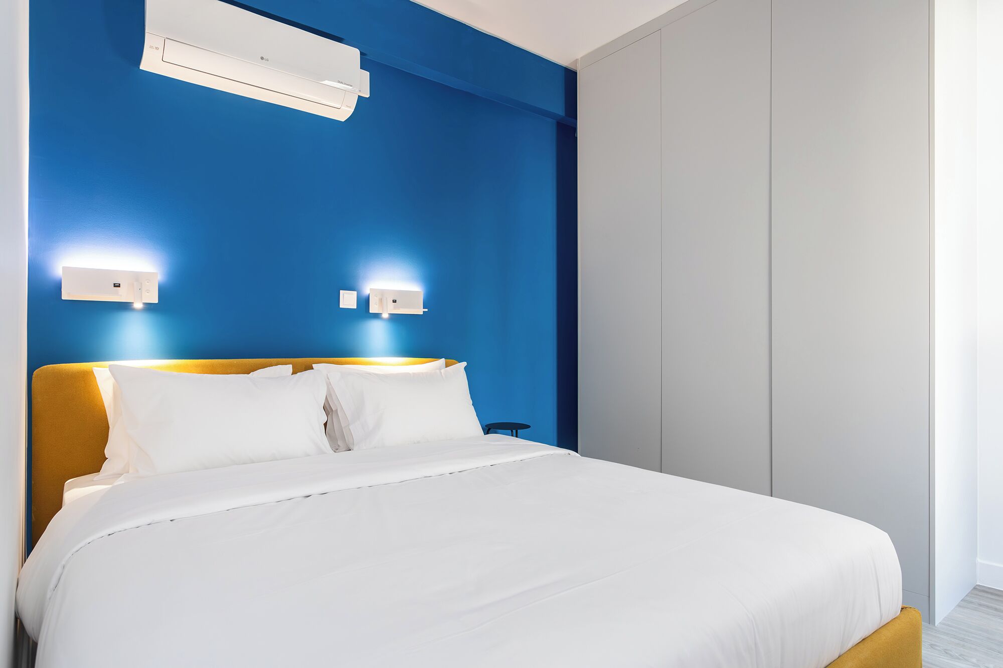 A hotel room with a bed, a wall-mounted air conditioner, and two wall-mounted light fixtures.