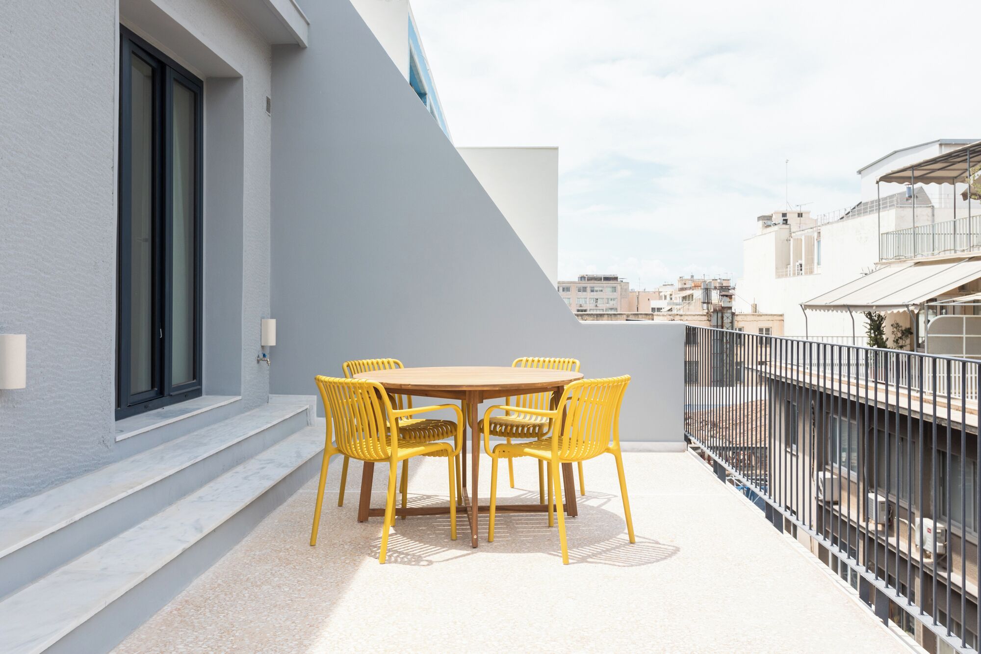 A balcony with a round wooden table and four yellow chairs, a white wall, and a metal railing.