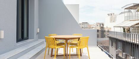 A balcony with a round wooden table and four yellow chairs, a white wall, and a metal railing.