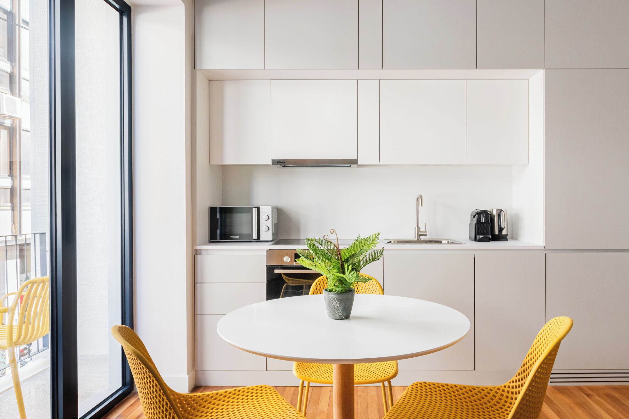 A modern kitchen with white cabinets, a microwave, a coffee maker, and a potted plant on the counter. There is a round white table with yellow chairs in front.