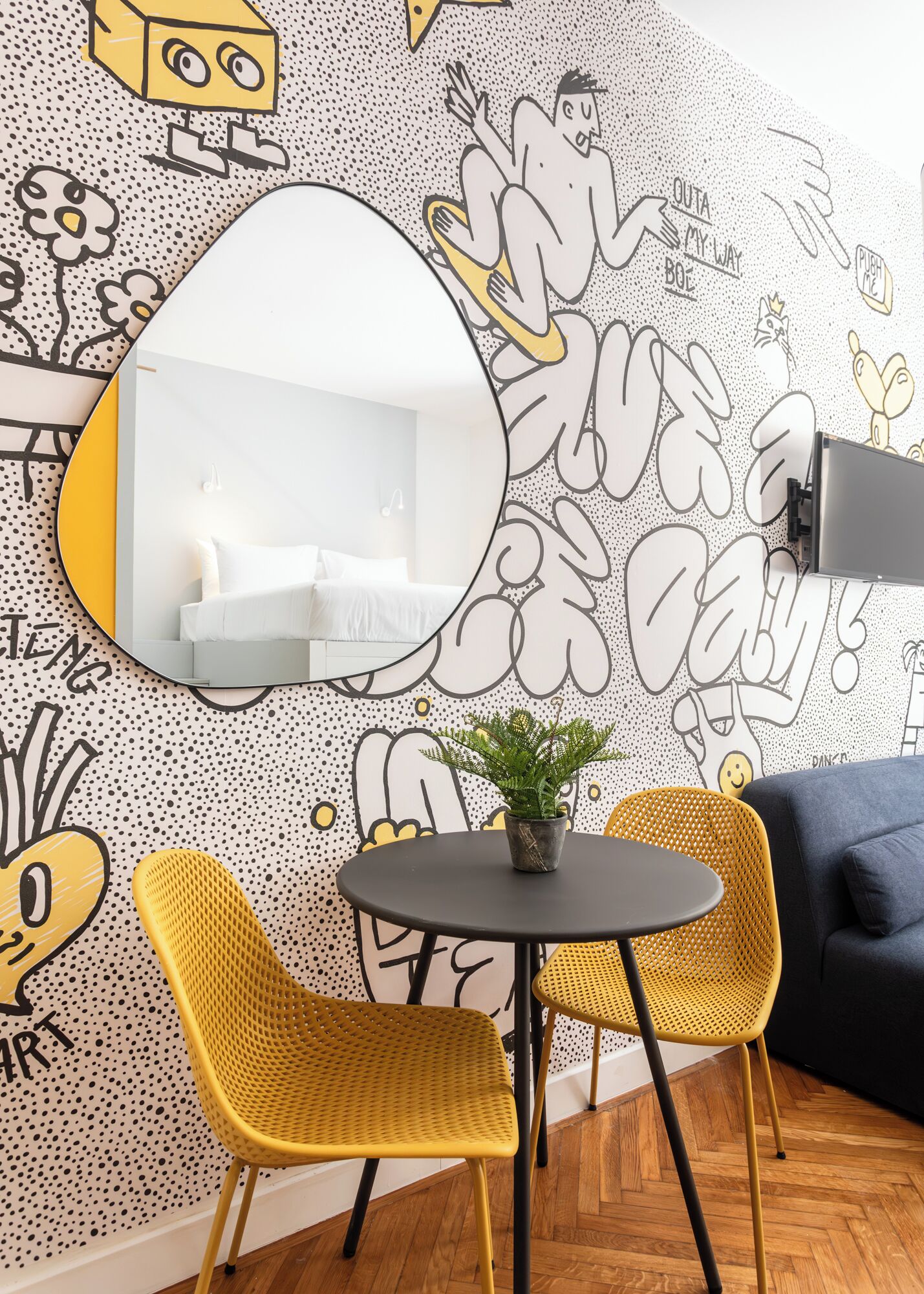 Un salon moderne avec un grand miroir, deux fauteuils en osier jaune, une petite table ronde agrémentée d’une plante et un canapé bleu.